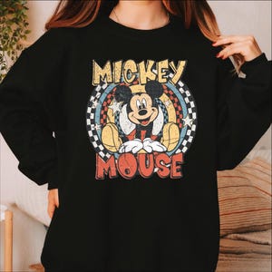 K&ouml;nnte beinhalten: Schwarzes Sweatshirt mit einem Retro-Mickey-Mouse-Motiv. Das Design zeigt die Worte "Mickey Mouse" in einer Vintage-Schriftart, mit Mickey in der Mitte. Die Grafik hat ein kreisf&ouml;rmiges Design mit Sternen und einem Schachbrettmuster.