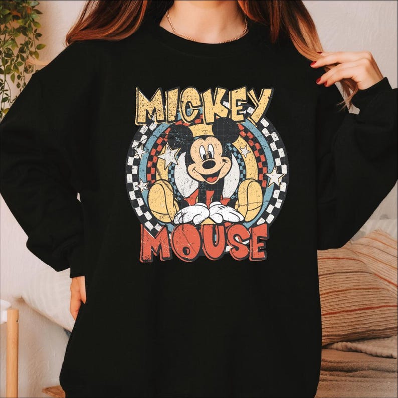 K&ouml;nnte beinhalten: Schwarzes Sweatshirt mit einem Retro-Mickey-Mouse-Motiv. Das Design zeigt die Worte "Mickey Mouse" in einer Vintage-Schriftart, mit Mickey in der Mitte. Die Grafik hat ein kreisf&ouml;rmiges Design mit Sternen und einem Schachbrettmuster.