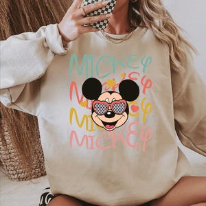 May include: Beige sweatshirt featuring a graphic of Mickey Mouse wearing heart-shaped sunglasses with a blue and white pattern. The word "MICKEY" is printed in multiple colors across the front.