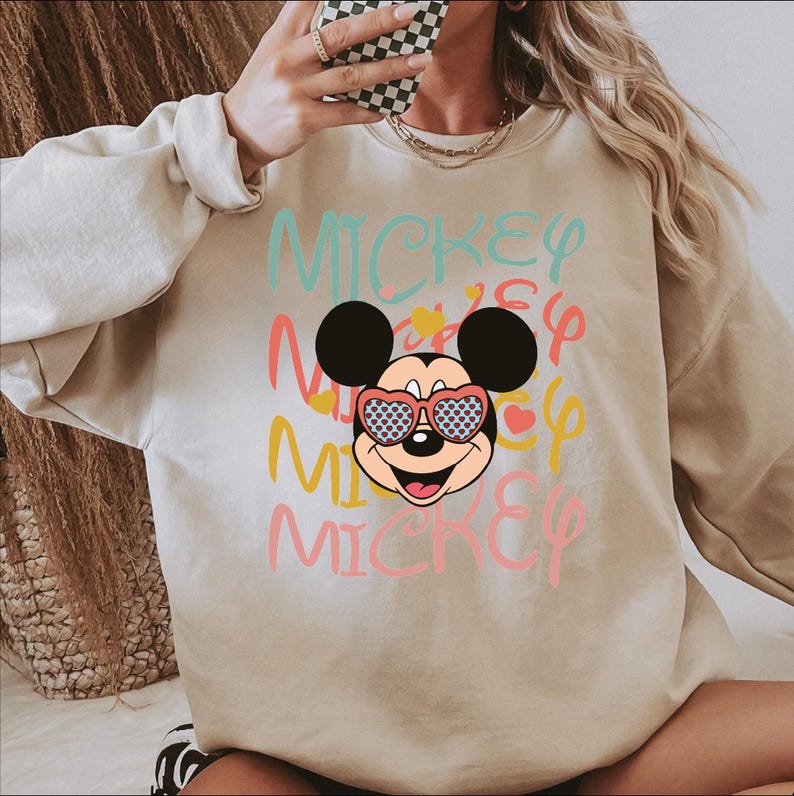 May include: Beige sweatshirt featuring a graphic of Mickey Mouse wearing heart-shaped sunglasses with a blue and white pattern. The word "MICKEY" is printed in multiple colors across the front.