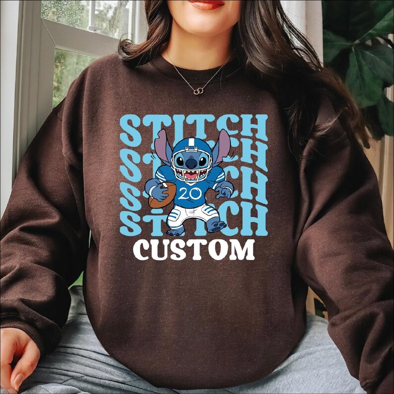 May include: A brown sweatshirt featuring a cartoon character in a football uniform. The character is blue with large ears and is holding a football. The word "STITCH" is printed in light blue, along with the word "CUSTOM" in white.