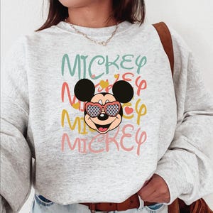 May include: A light gray sweatshirt featuring a graphic of Mickey Mouse wearing heart-shaped sunglasses. The word "MICKEY" is repeated in various colors, including teal, yellow, and pink, creating a playful design.