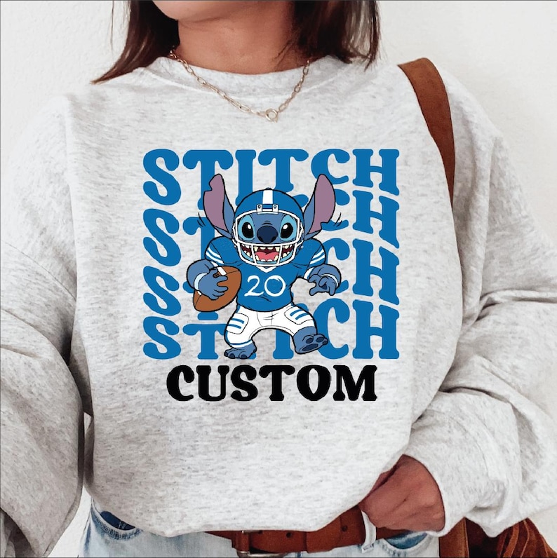 May include: A light gray sweatshirt featuring a blue cartoon character in a football uniform holding a football. The word "STITCH" is repeated in blue, with "CUSTOM" in black below the character.