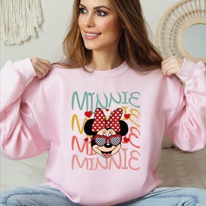 Puede incluir: Sudadera rosa claro con un dise&ntilde;o de Minnie Mouse. El dise&ntilde;o incluye un lazo rojo con lunares, gafas de sol y la palabra "MINNIE" en varios colores. La sudadera es de cuello redondo.