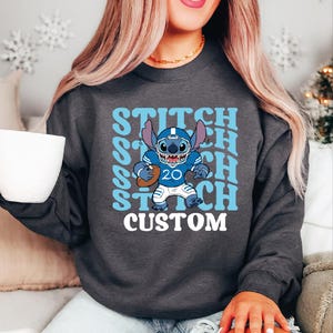 May include: Dark gray sweatshirt featuring a cartoon Stitch character in a football uniform. The design includes the word "STITCH" in light blue, repeated vertically, and the word "CUSTOM" in white.