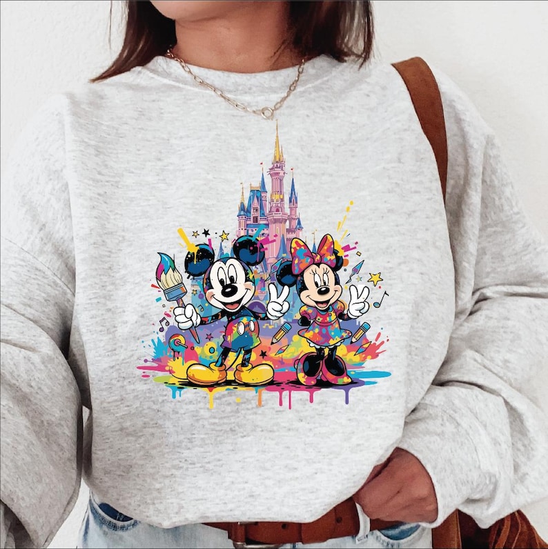 Peut inclure: Sweat-shirt gris clair avec un motif color&eacute; de Mickey et Minnie Mouse devant un ch&acirc;teau. Mickey tient un pinceau, Minnie fait le signe de la paix, et tous deux sont entour&eacute;s d'&eacute;claboussures de peinture et de notes de musique.
