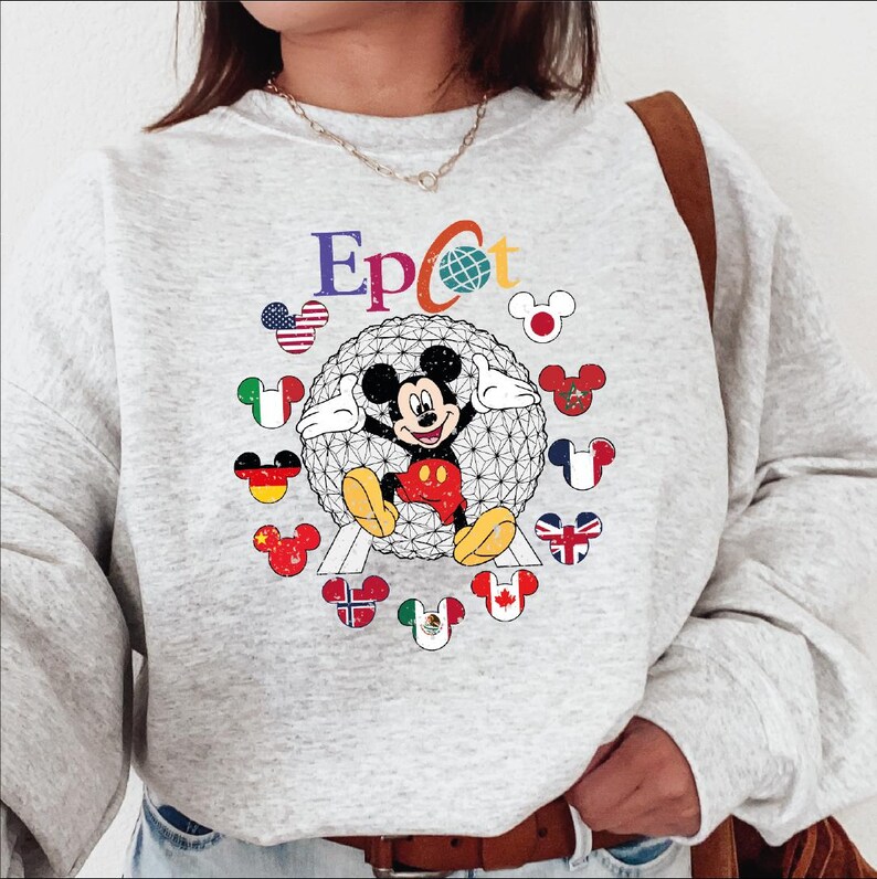 May include: A light gray sweatshirt featuring the word "Epcot" in colorful letters, with a globe graphic. Mickey Mouse is in the center, surrounded by Mickey Mouse heads with flags. The sweatshirt has long sleeves and a crew neck.