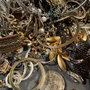 May include: A close-up shot of a collection of mixed jewelry, including necklaces, bracelets, and rings. The jewelry pieces are in various colors, including gold, silver, and bronze. Some pieces feature pearls and gemstones. A coin is also visible.
