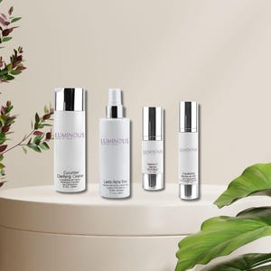 Könnte beinhalten: Vier weiße Hautpflegeproduktflaschen mit silbernen Kappen sind auf einem weißen Podest angeordnet. Die Flaschen sind mit "Cucumber Clarifying Cleanse", "Lactic Alpha Toner", "Radian-C Serum" und "Equalizing Moisture Gel" beschriftet. Die Marke "LUMINUS" ist sichtbar.