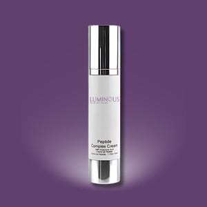 Puede incluir: Un frasco de crema cosmética blanco y plateado sobre un fondo púrpura. El frasco tiene el texto "LUMINOUS SKIN BY JAIRE" y "Peptide Complex Cream" impreso. El frasco es cilíndrico con tapa y base plateadas.