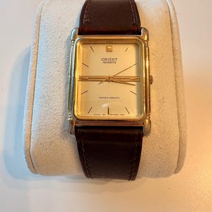 Puede incluir: Un reloj Orient Quartz dorado con una esfera rectangular y una correa de cuero marrón. La esfera del reloj es de color oro claro con marcas y agujas negras. El reloj se muestra sobre un cojín de color crema.