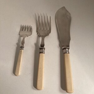 Può includere: Un set di posate antiche composto da due forchette e un coltello da burro. Le forchette hanno più rebbi e manici color crema. Il coltello da burro ha una lama larga e curva e un manico abbinato. Le posate sono in metallo.