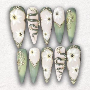 May include: A set of ten long, almond-shaped artificial nails. The nails feature a variety of designs, including white floral patterns, gold accents, and a gold snake. The color palette includes pale pink, green, and gold.