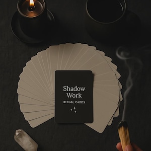 Può includere: Un'immagine still life con un mazzo di carte rituali beige con una carta nera che recita "Shadow Work RITUAL CARDS". Sono presenti anche una candela accesa, cristalli e un bastoncino di fumo fumante, creando un'estetica mistica.