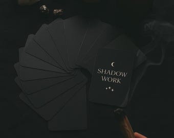 May include: A still-life arrangement featuring a deck of black cards fanned out, with one card displaying the text "SHADOW WORK". A lit candle, a cup, crystals, and a burning stick are also present, creating a mystical ambiance.