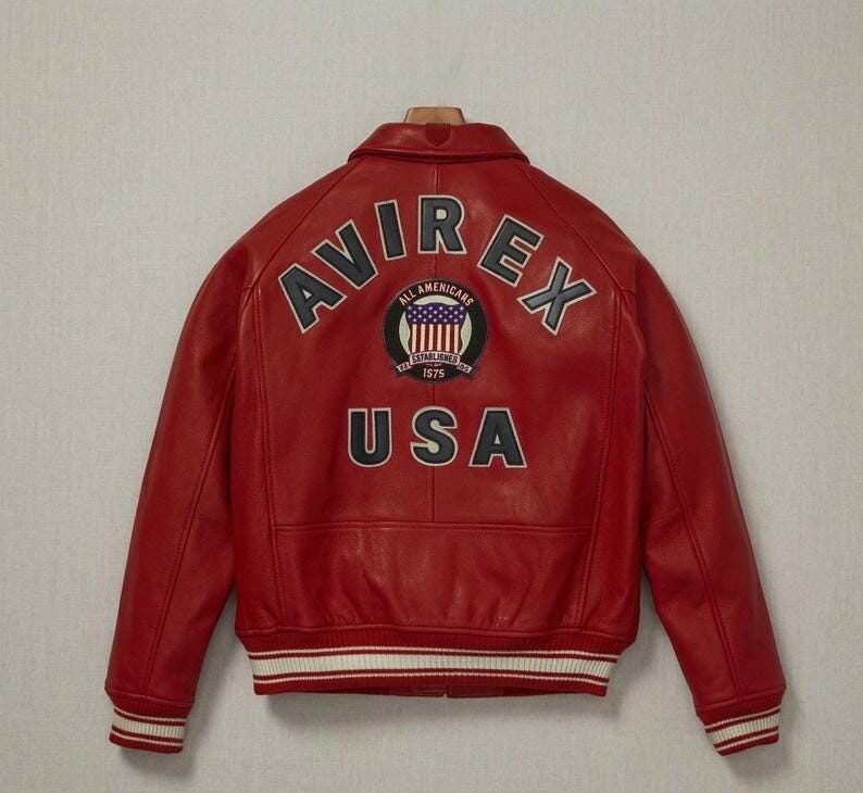 May include: A bright red leather jacket featuring a fold-down collar, long sleeves, and ribbed cuffs and hem with white stripes. The back displays the text AVIREX in an arc, a circular patch with a flag graphic, and the text USA in large letters.