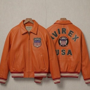 May include: An orange leather jacket with a front zipper, a collar, and striped ribbed cuffs and hem. The front features an Avirex shield patch. The back displays the text Avirex and USA with a circular patch containing a flag and the text 1975.