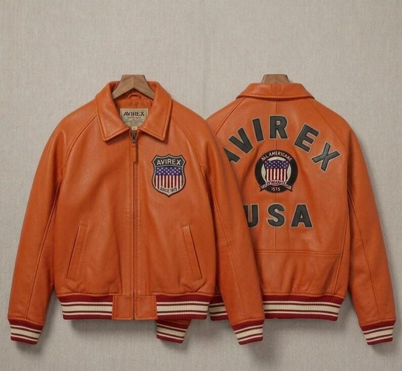 May include: An orange leather jacket with a front zipper, a collar, and striped ribbed cuffs and hem. The front features an Avirex shield patch. The back displays the text Avirex and USA with a circular patch containing a flag and the text 1975.