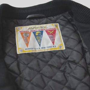 Op de afbeelding: Een zwarte jas met een doorgestikte binnenvoering en een rechthoekig wit label. Het label bevat de tekst AVIREX AUTHENTIC VARSITY SPORTS EQUIPMENT en AVIREX U.S.A. NEW YORK N.Y. met drie kleurrijke wimpelafbeeldingen.