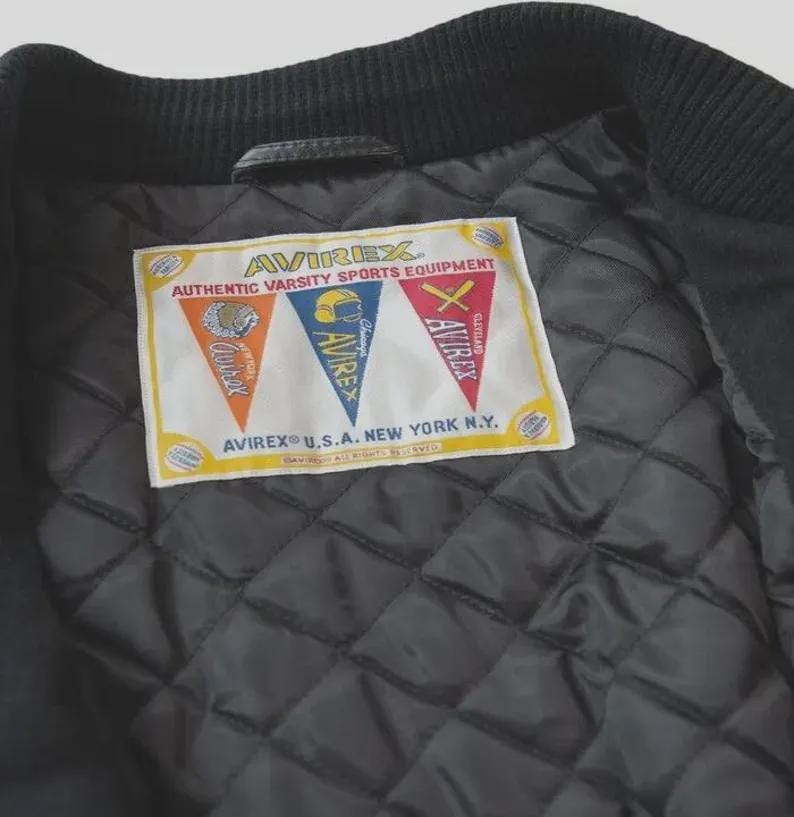 Op de afbeelding: Een zwarte jas met een doorgestikte binnenvoering en een rechthoekig wit label. Het label bevat de tekst AVIREX AUTHENTIC VARSITY SPORTS EQUIPMENT en AVIREX U.S.A. NEW YORK N.Y. met drie kleurrijke wimpelafbeeldingen.