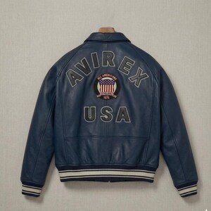 May include: A navy blue leather bomber jacket featuring the text AVIREX arched across the upper back in grey lettering. Below the text is a circular patch with a flag design and the word USA in large grey letters at the bottom hem.