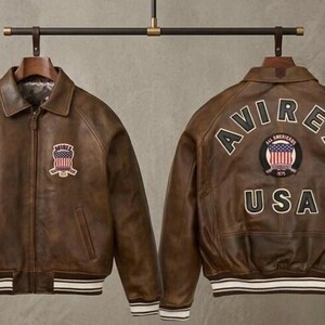 May include: A brown leather bomber jacket shown from the front and back. The front features a small patch with a flag design and the text AVIREX. The back displays the text AVIREX, a circular flag patch, and the text USA. It has striped ribbed cuffs.