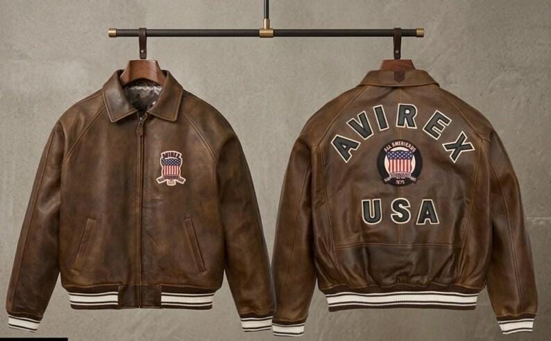 May include: A brown leather bomber jacket shown from the front and back. The front features a small patch with a flag design and the text AVIREX. The back displays the text AVIREX, a circular flag patch, and the text USA. It has striped ribbed cuffs.