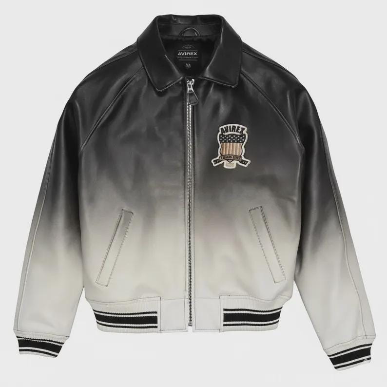 May include: A black and white ombre leather jacket with a full-length front zipper, a classic collar, and two side pockets. The chest features an embroidered patch with the text AVIREX and a shield graphic containing a star-spangled banner design.
