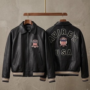 May include: A black leather jacket with a front zipper, a collar, and striped ribbed cuffs and hem. The front features an Avirex patch with a flag design. The back displays the text Avirex and USA with a circular patch featuring a flag and text.