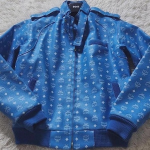 May include: A bright blue jacket featuring a repeating white MCM logo and diamond pattern. The jacket has a full-length front zipper, a stand-up collar with a BOSS brand tag, long sleeves with ribbed cuffs, and a ribbed hem. The material has a textured finish.