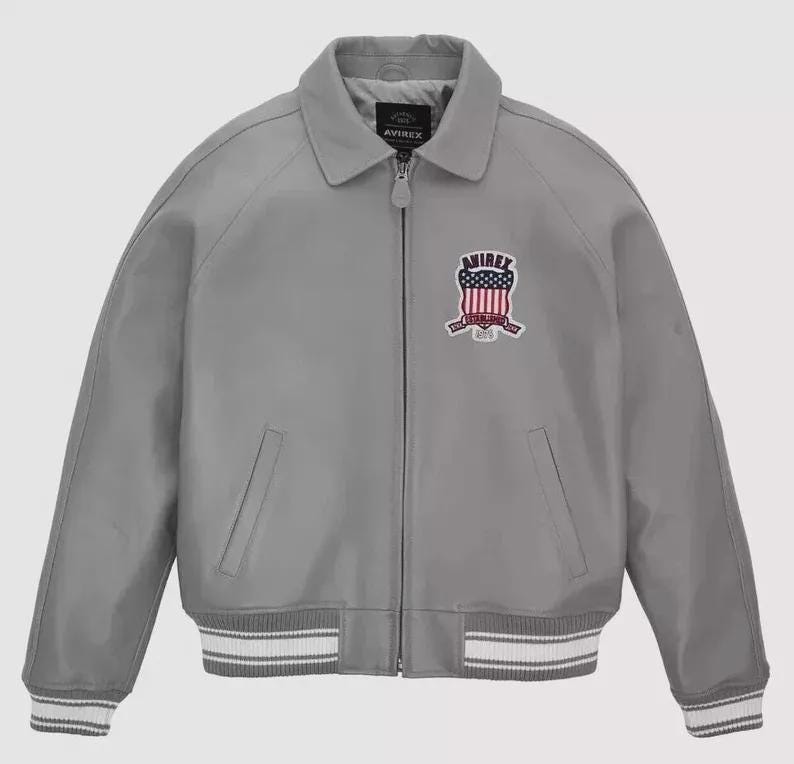 Może przedstawiać: Szara kurtka bomberka z kołnierzem i zapięciem na zamek błyskawiczny. Kurtka posiada naszywkę z czerwono-biało-niebieskim emblematem. Mankiety i d&oacute;ł mają białe paski. Widoczna jest nazwa marki "AVIREX".