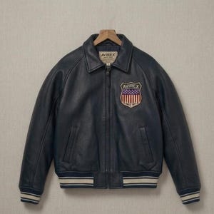 May include: A dark navy blue leather bomber jacket with a full-length front zipper, a pointed collar, and ribbed cuffs and hem featuring white and navy stripes. A shield-shaped patch on the chest displays the American flag and the text AVIREX.