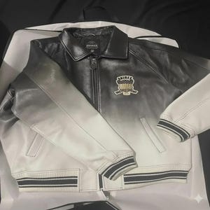 May include: A black and white leather jacket featuring a gradient fade from dark at the collar to light at the hem. The jacket has a front zipper, a pointed collar, ribbed cuffs and hem with black stripes, and an Avirex patch on the chest.