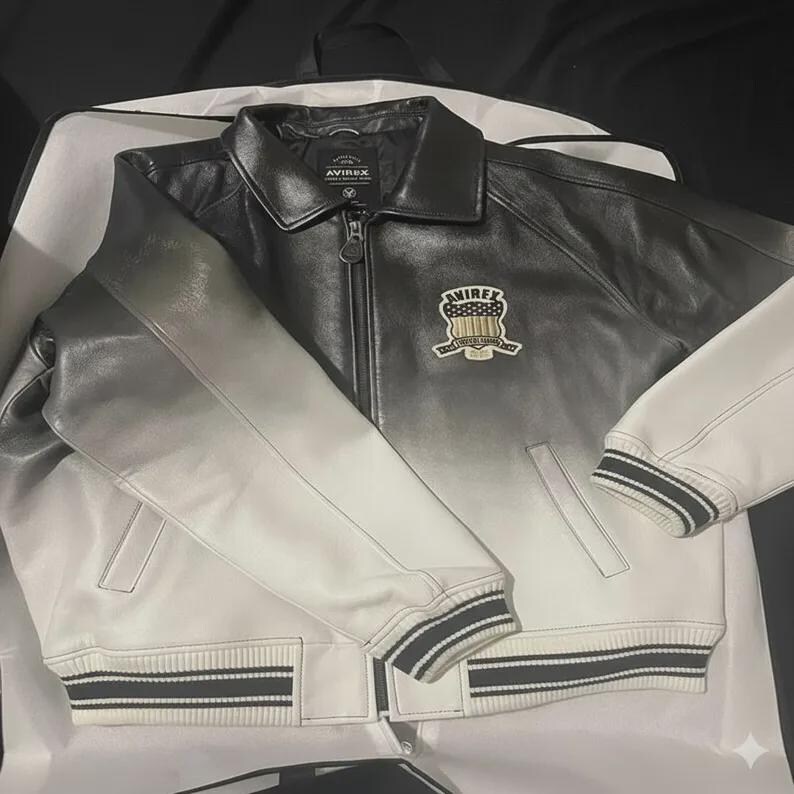 May include: A black and white leather jacket featuring a gradient fade from dark at the collar to light at the hem. The jacket has a front zipper, a pointed collar, ribbed cuffs and hem with black stripes, and an Avirex patch on the chest.