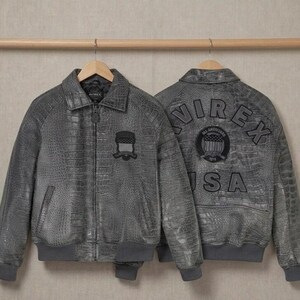 May include: Two grey jackets with a textured crocodile skin pattern. The front features a collar, zipper, and a small shield patch. The back displays the text AVIREX USA in large letters and a circular patch with a flag design and text.