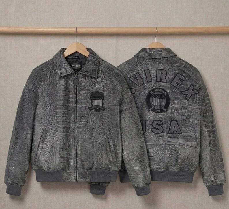 May include: Two grey jackets with a textured crocodile skin pattern. The front features a collar, zipper, and a small shield patch. The back displays the text AVIREX USA in large letters and a circular patch with a flag design and text.
