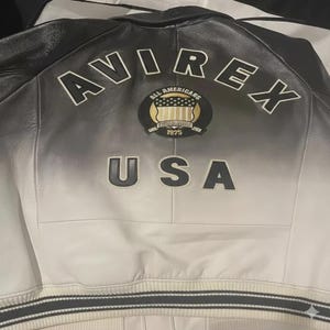 May include: A black and white leather jacket featuring a gradient fade design. The back displays the text AVIREX in an arc, a circular patch with the text ALL AMERICANS and 1975, and the text USA below. The jacket has a ribbed hem with stripes.