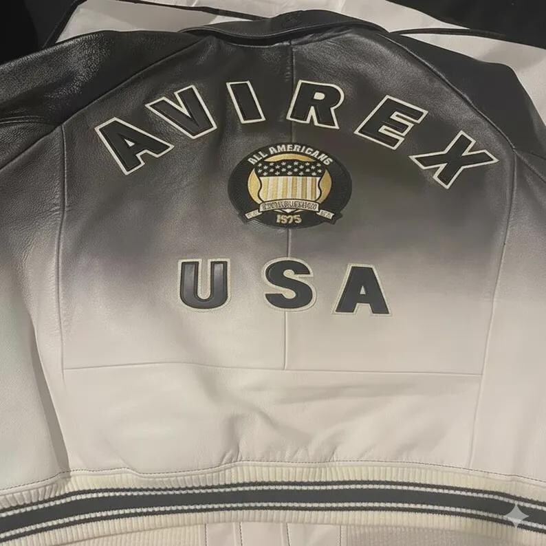 May include: A black and white leather jacket featuring a gradient fade design. The back displays the text AVIREX in an arc, a circular patch with the text ALL AMERICANS and 1975, and the text USA below. The jacket has a ribbed hem with stripes.