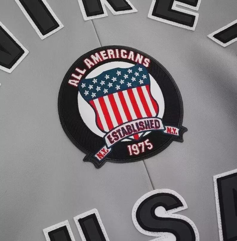 Może przedstawiać: Zbliżenie szarej kurtki z okrągłą naszywką. Naszywka przedstawia wz&oacute;r tarczy z czerwono-białymi paskami i niebieskimi gwiazdami, ze słowami "ALL AMERICANS" i "ESTABLISHED 1975 N.Y." w czarnej obw&oacute;dce. Czarne i białe litery są rozrzucone wok&oacute;ł naszywki.