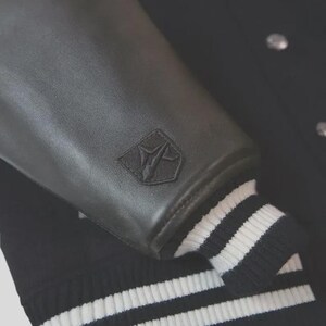 Op de afbeelding: Een zwart varsity-jack met gladde zwarte leren mouwen en een zwarte wollen body. Op de mouw bevindt zich een klein, tonaal geborduurd schildvormig logo. De boorden aan de mouwen en de onderkant zijn afgewerkt met zwart-wit gestreept ribgebreid materiaal.
