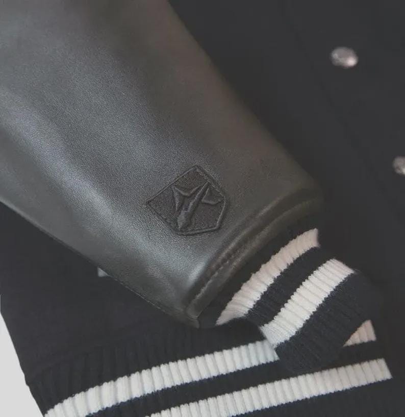 Op de afbeelding: Een zwart varsity-jack met gladde zwarte leren mouwen en een zwarte wollen body. Op de mouw bevindt zich een klein, tonaal geborduurd schildvormig logo. De boorden aan de mouwen en de onderkant zijn afgewerkt met zwart-wit gestreept ribgebreid materiaal.