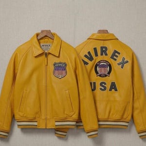 May include: A bright yellow leather bomber jacket featuring a front zipper, a collar, and striped ribbed cuffs and hem. The front displays an Avirex shield patch. The back features the text Avirex and USA with a circular All American shield patch.