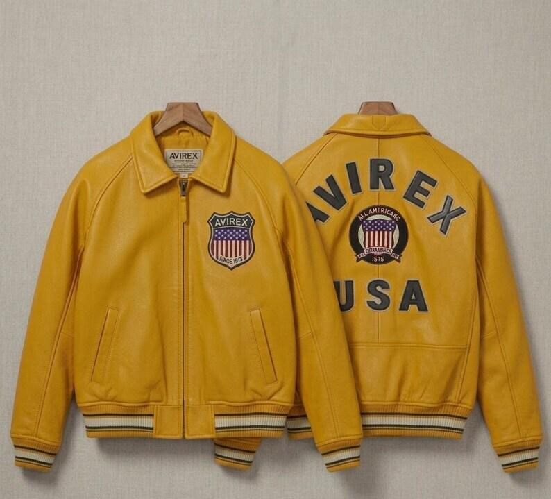May include: A bright yellow leather bomber jacket featuring a front zipper, a collar, and striped ribbed cuffs and hem. The front displays an Avirex shield patch. The back features the text Avirex and USA with a circular All American shield patch.