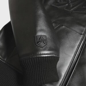 May include: Black leather jacket with a ribbed cuff and a shield-shaped emblem on the sleeve. The jacket features a zipper closure and embossed detailing on the upper back. The overall design is sleek and modern.