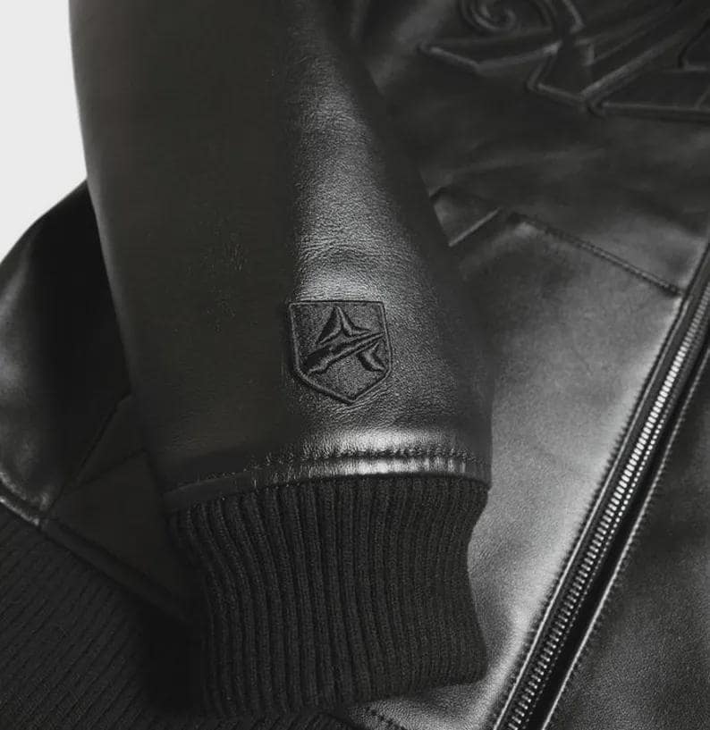 May include: Black leather jacket with a ribbed cuff and a shield-shaped emblem on the sleeve. The jacket features a zipper closure and embossed detailing on the upper back. The overall design is sleek and modern.