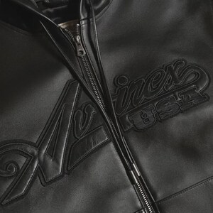 May include: Black leather jacket with a full zipper closure. The jacket features the word "Avirex" in a stylized font, with "USA" underneath. The jacket has a classic bomber style.