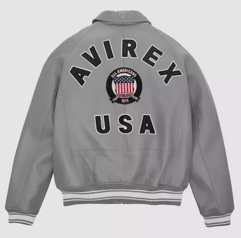 Może przedstawiać: Szara kurtka bomberka z napisem "AVIREX" wygiętym na plecach, nad okrągłym emblematem i napisem "USA". Emblemat przedstawia tarczę z czerwono-białymi paskami i niebieskimi gwiazdami. Kurtka ma biało-szare paski na mankietach.