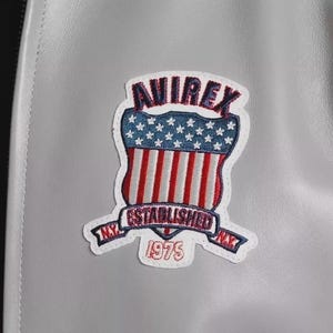 Może przedstawiać: Zbliżenie szarej sk&oacute;rzanej kurtki z kolorową naszywką. Na naszywce widnieje napis "AVIREX" w kolorze czerwonym, niebiesko-biała tarcza z gwiazdami, czerwono-białe paski oraz napisy "ESTABLISHED NY 1975".