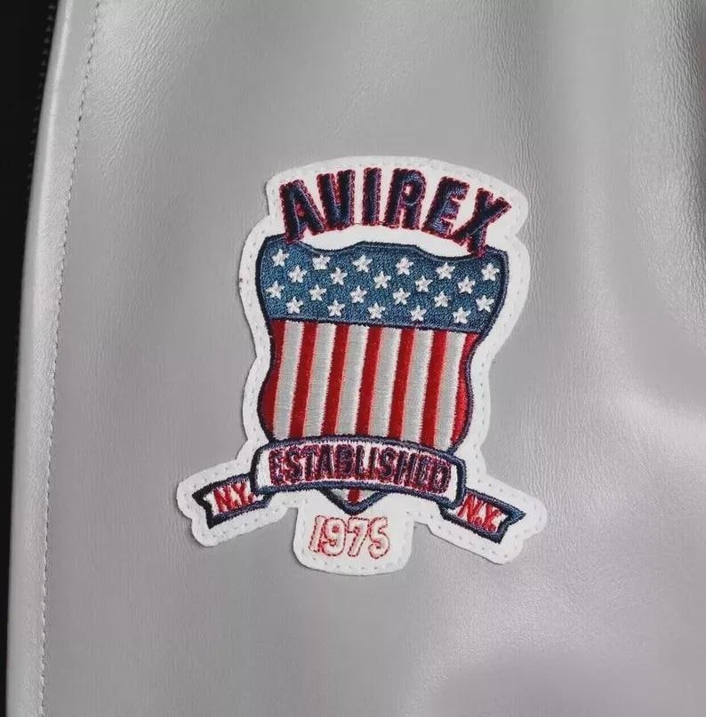 Może przedstawiać: Zbliżenie szarej sk&oacute;rzanej kurtki z kolorową naszywką. Na naszywce widnieje napis "AVIREX" w kolorze czerwonym, niebiesko-biała tarcza z gwiazdami, czerwono-białe paski oraz napisy "ESTABLISHED NY 1975".