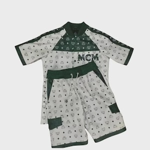 Könnte beinhalten: Ein zweiteiliges Set bestehend aus einem kurzärmeligen Hemd und Shorts. Das Set ist weiß mit einem sich wiederholenden schwarzen Muster und dunkelgrünen Akzenten. Auf dem Hemd und den Shorts sind die Buchstaben "MCM" aufgedruckt.
